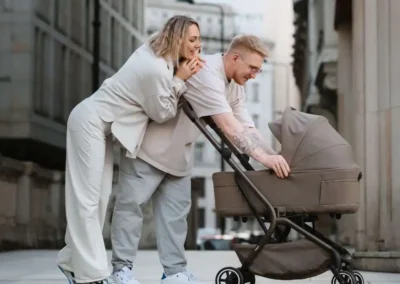 Couple pushing a beige baby stroller on a city sidewalk, leaning in to look at the baby inside the stroller.